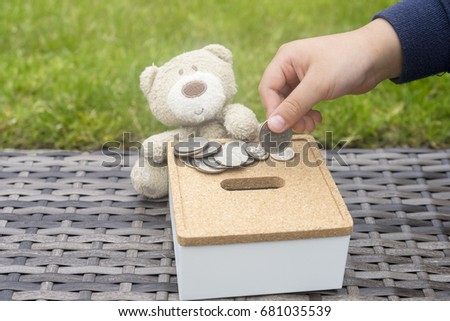 Close up kid hand putting coin in the white square box with teddy bear sitting on rattan, Saving money concept