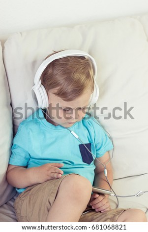 3 year old boy sits on the couch and listens to music. Little cute boy watching cartoons on the phone. Little boy.