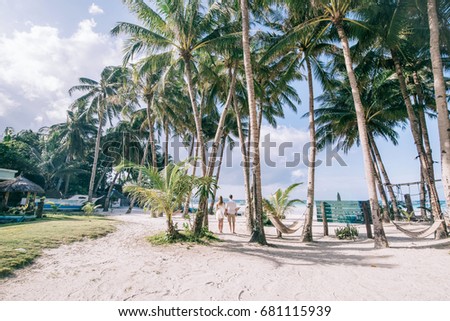 Young couple look to the sea,the fresh green palm trees around. They stand on the white sand with a tropical sea and blue sky with clouds on the background. 
