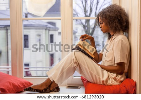 Slender african american girl sitting on the windowsill with the book in her hands.