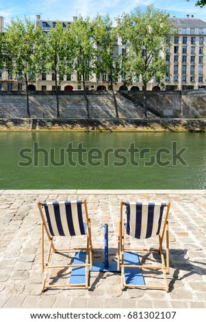 Two blue and white striped deck chairs in the sun on the bank of the river Seine with poplar trees and typical parisian buildings in the background.
