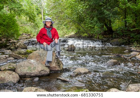 Smiling woman sitting on rock by river, small waterfall in background