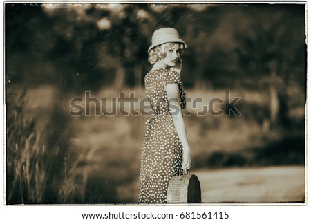 Classic black and white photo of 1930s summer fashion woman with dress and straw hat standing with handbag in rural landscape. Looking over shoulder.
