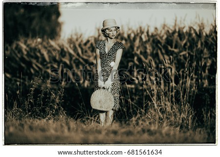 Classic black and white photo of 1920s summer fashion woman with dress and straw hat standing with handbag in rural landscape.