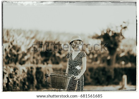 Classic black and white photo of 1930s fashion woman in summer dress standing with bicycle in rural landscape.