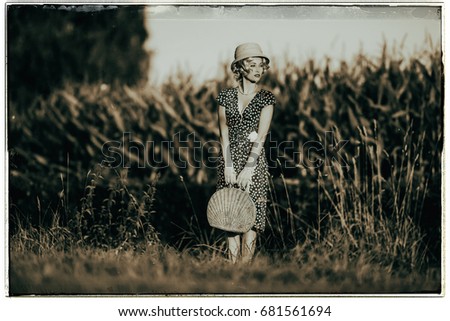 Classic black and white photo of 1920s summer fashion woman with dress and straw hat standing with handbag in rural landscape.