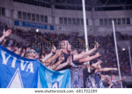 Blurred background of crazy football fans at the stadium. Fanatical fans in stands during game of rivals. Fans in stands rejoice, shout and fluttering flag. People are looking at stadium. Strong blur