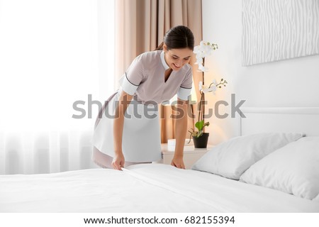 Young maid making bed in light hotel room Royalty-Free Stock Photo #682155394