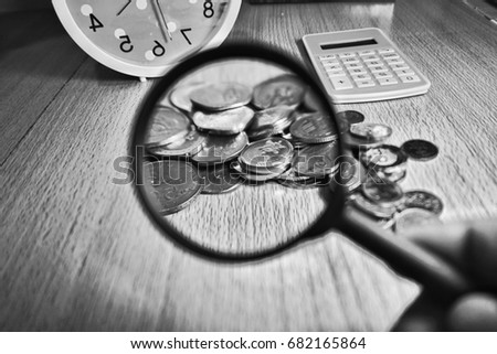Black and white conceptual photo including calculator, coins, wooden background and magnifying glass to show financial related issues. Selective Focused.