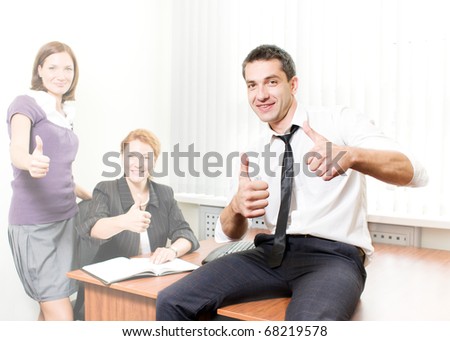 Smiling member of business team showing two thumb-up together with his office workers. Handsome man in business suit sitting on table and smiling for camera.
