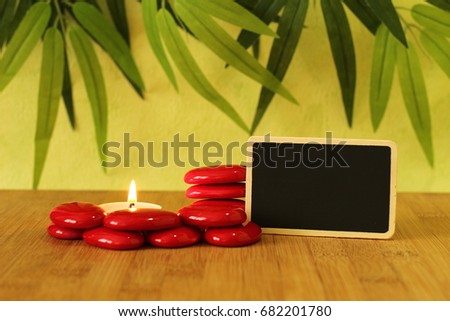 Small empty slate in width to write a message posed on the bamboo floor with red stones columns in the lifestyle Zen with a candle all on wooden floor and green foliage background