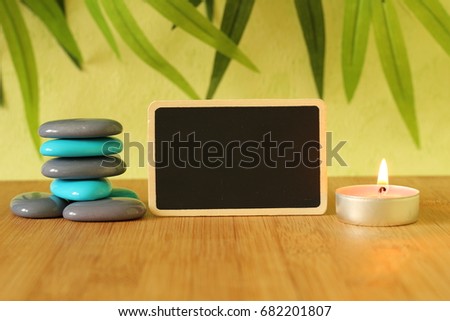Small empty slate in width to write a message posed on the bamboo floor with gray and blue stones columns in the lifestyle Zen with a candle all on wooden floor and green foliage background