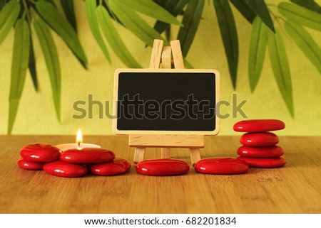 Small empty slate in width to write a message that is posed on an easel with red stones  columns on the ground in zen lifestyle with a candle all on wooden floor and green foliage background