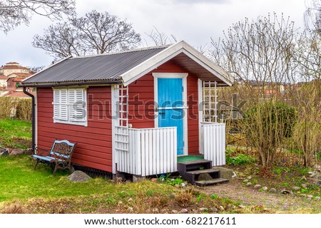 Red tiny house or shed at a plot of an allotment. Early spring time. Small red shack with blue door, white frames, shutters and metal forged bench by wall. Hut painted in traditional Swedish red color Royalty-Free Stock Photo #682217611