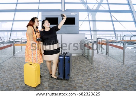 Two fat women taking a selfie photo by using smartphone while standing with a suitcase in the airport terminal