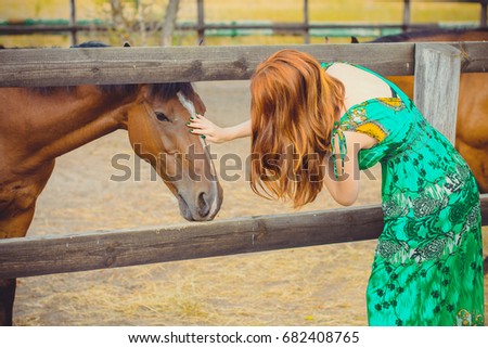 Woman with red hair and ornament green ethnic dress and with horses on a farm, pets animals in village in a rancho. Horses are human friends