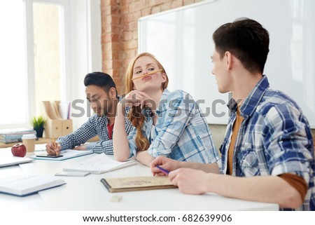 Funny girl with pencil between her upper lip and nose looking at one of her groupmates in classroom