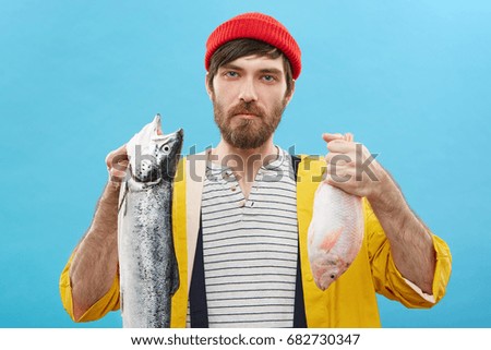 Picture of handsome young Caucasian angler or fisherman with blue eyes and beard holding two fish, looking grumpy and dissatisfied, unhappy with poor catch. Hobby, leisure and occupation concept