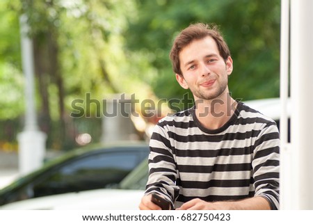 Smiling hipster guy holding mobile phone and looking at the camera while waiting for someone outdoors,cheerful man wanderer use cell telephone while standing near table with copy space area