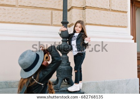 Cute little girl with funny face expression standing near the iron pillar, posing in front of mother in trendy felt hat. Young woman with professional camera making photoshoot for daughter.