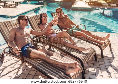 Group of young friends is lying on a chaise-longues near swimming pool and smiling. Having rest and drinking beer together.
