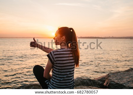A young girl sits on the rocks next to the sea at sunset and shows a hand sign which means cool. Rest, vacation, relaxation.