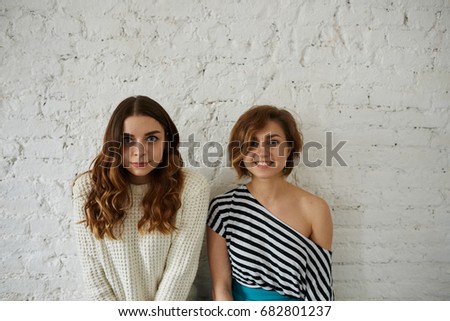 Indoor fashion portrait of two charming young women best friends, wearing stylish clothes and natural make-up, posing for picture against white brick wall background with copy space for your text