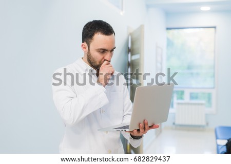 Minded successful doctor standing at his office and using laptop for his work