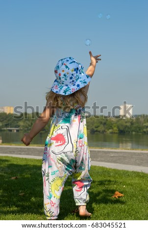 Cute little girl in a hat jumps on the green lawn next to river and stretches her hands to the soap bubbles of the flying in front of her. Sunny morning in the city park. Back view.