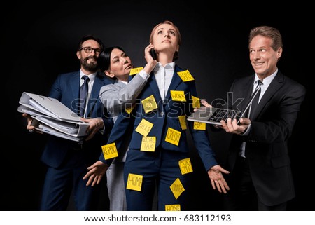 stressed mature businesswoman with sticky notes on clothes standing between colleagues holding folders, smartphone and laptop isolated on black