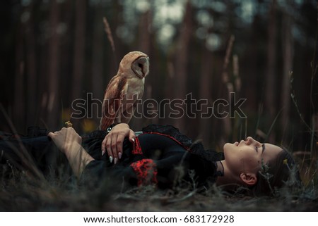 Girl in red and black dress lies with owl on grass in forest. Owl sits on her hand.