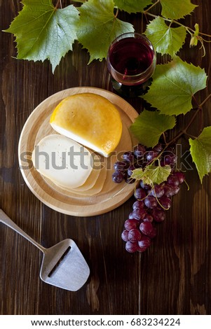Small round head of cheese, cheese knife, grapes and leaves of vine on rustic wooden background