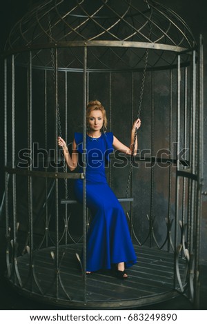 Beautiful girl with blond hair sitting on a swing in a long blue evening dress in an interior studio. Fashion shooting