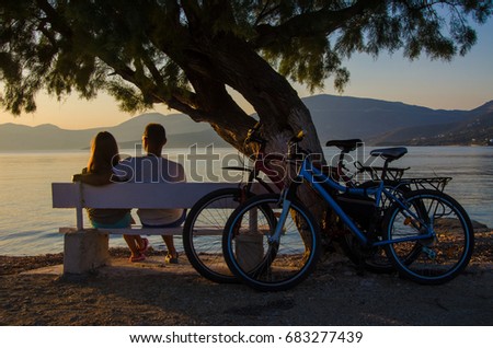 a couple of people sitting on a bench under a tree by the sea while watching the sunset, having arrived on a bicycle