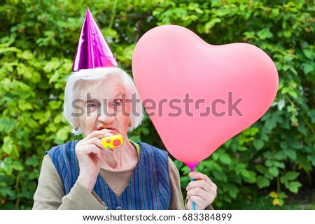 Picture of a happy elderly woman celebrating birthday holding a heart shaped balloon an blowing a whistle outdoor