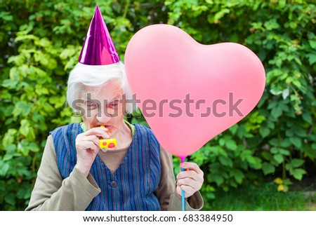 Picture of a happy elderly woman celebrating birthday holding a heart shaped balloon an blowing a whistle outdoor