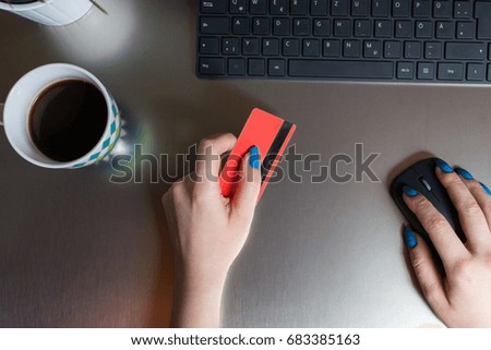 Cropped top view image of female hand holding credit card and computer mouse while sitting at office table. Cup of coffee and a green plant aside. Doing some online shopping