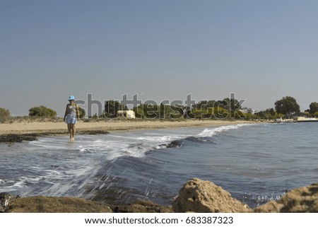 Women in hat walking barefoot at sea shore with blue sky in background and wave from the sea in foreground, picture from Northern Cyprus.