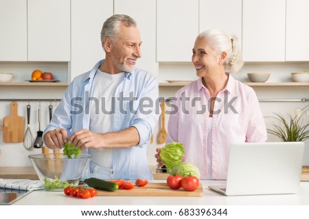 Picture of mature loving couple family standing at the kitchen using laptop computer and cooking salad. Looking aside.