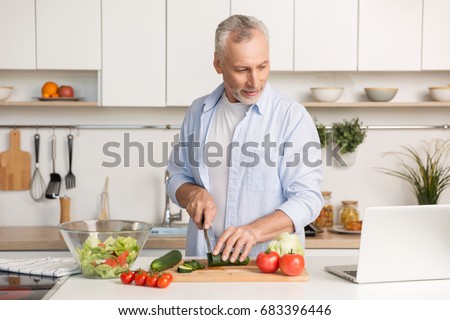 Picture of mature handsome man standing at the kitchen using laptop computer and cooking salad. Looking aside.