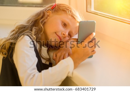 A happy european girl with a mobile phone indoors, near a window, looking at the screen with interest, watching cartoons or playing video games.
After school hours