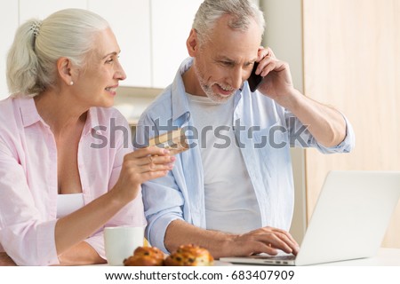 Picture of smiling mature loving couple family standing at the kitchen using laptop computer holding credit card. Looking aside talking by phone.