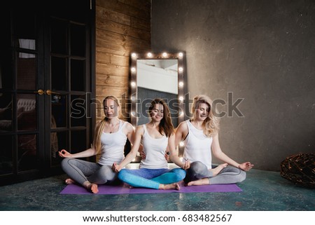 Three beautiful girls sit side by side in lotus position in yoga classroom