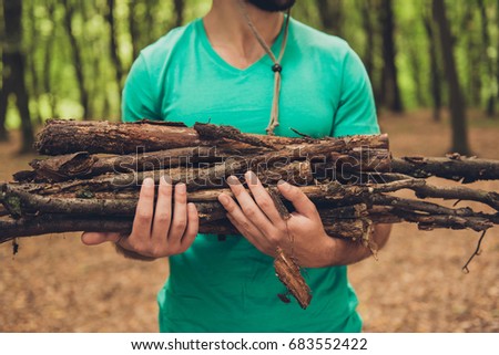 Cropped close up photo of a bearded male tourist during hiking, holding wood for a campfire, in green t shirt, in a forest in the early fall Royalty-Free Stock Photo #683552422