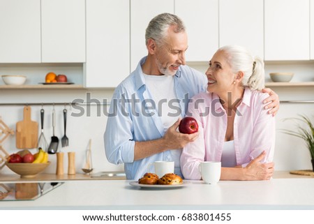 Picture of smiling mature loving couple family standing at the kitchen. Man holding apple. Looking at each other.