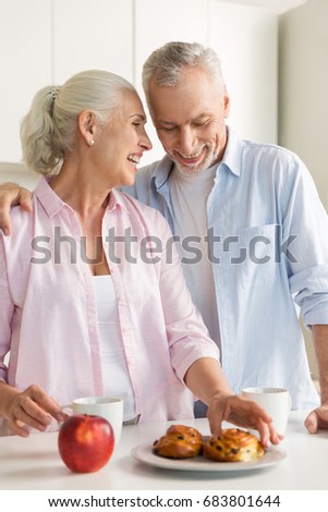 Photo of smiling mature loving couple family standing at the kitchen near apple and pastries. Looking aside.