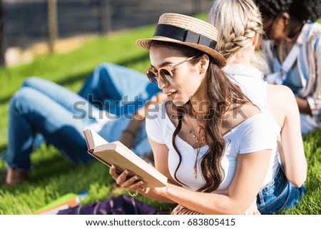 smiling young asian woman in sunglasses and straw hat reading book while sitting on grass with classmates talking behind