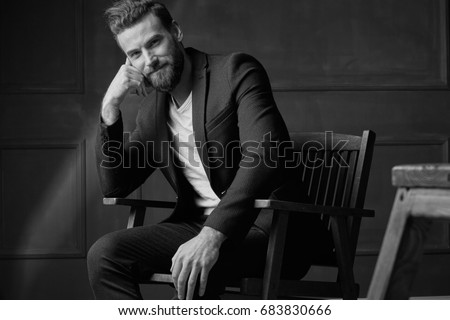 Handsome young beautiful and confident bearded man, wearing white shirt and brown jacket, sitting on wooden chair in studio with dark background and smiling, in black and white Royalty-Free Stock Photo #683830666