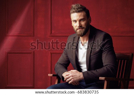 Handsome beautiful bearded man, wearing white shirt and brown jacket, sitting on wooden chair in studio with dark red background  Royalty-Free Stock Photo #683830735