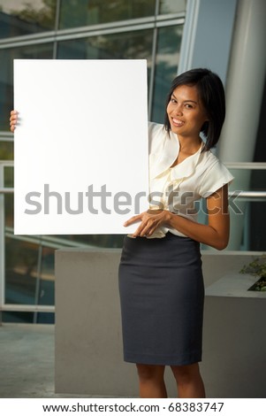 Attractive, smiling Asian business woman in white blouse holds a blank sign outside in front of a modern office complex.  Custom text copy space insert. Female Asian dark skin model Chinese descent